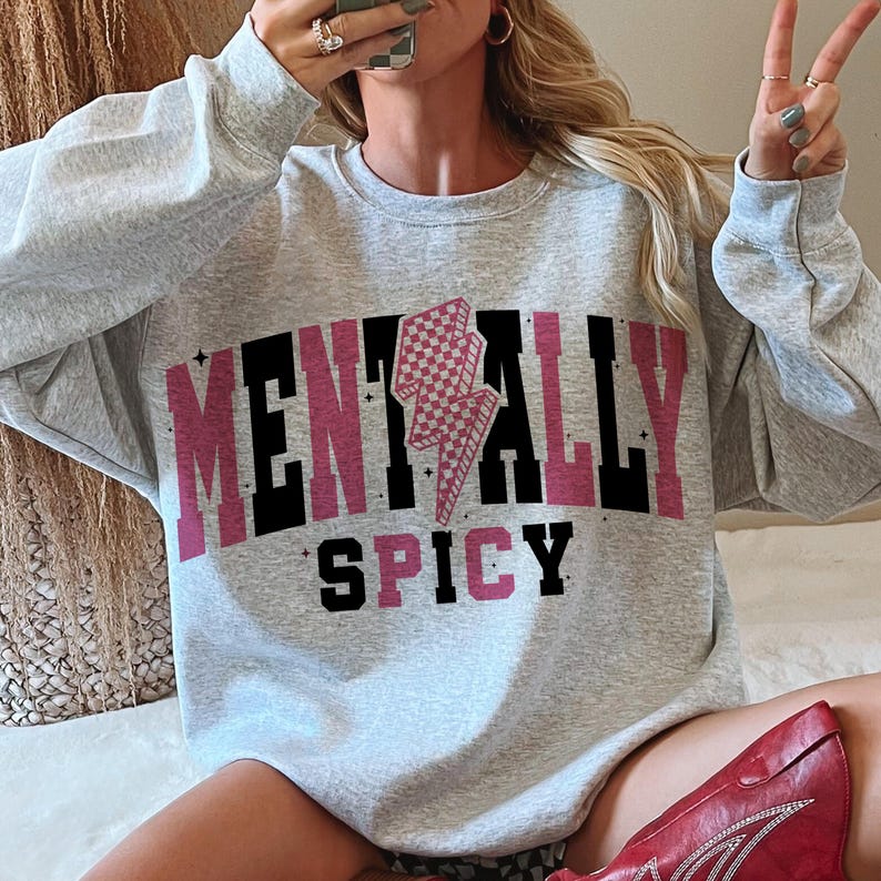 May include: A light gray sweatshirt with the words "MENTALLY SPICY" in a pink and black font. The letter "A" is replaced with a pink and white checkered lightning bolt graphic. The sweatshirt is a crew neck style.
