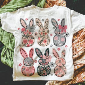 May include: White t-shirt with a floral pattern of six bunnies wearing bows. The bunnies are pink, gray, and green.