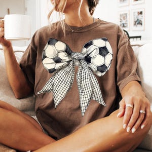Puede incluir: Una camiseta marr&oacute;n con un gr&aacute;fico de un lazo de bal&oacute;n de f&uacute;tbol. El lazo est&aacute; hecho de dos balones de f&uacute;tbol atados juntos con una cinta blanca.