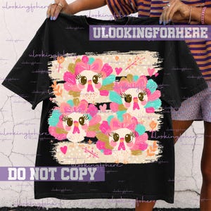 May include: A black short-sleeve t-shirt featuring a graphic print of four stylized turkeys with pink, teal, and gold feathers. The turkeys have large eyes, long eyelashes, and pink bows. The design includes small pink hearts and orange floral accents.
