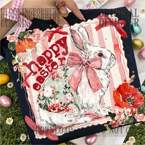 K&ouml;nnte beinhalten: Schwarzes T-Shirt mit einer Illustration eines wei&szlig;en Hasen mit rosa Schleife und Blumenmusterkleid. Die Worte "Happy Easter" sind rot. Das Design ist von Blumen umgeben, ideal f&uuml;r Ostern.