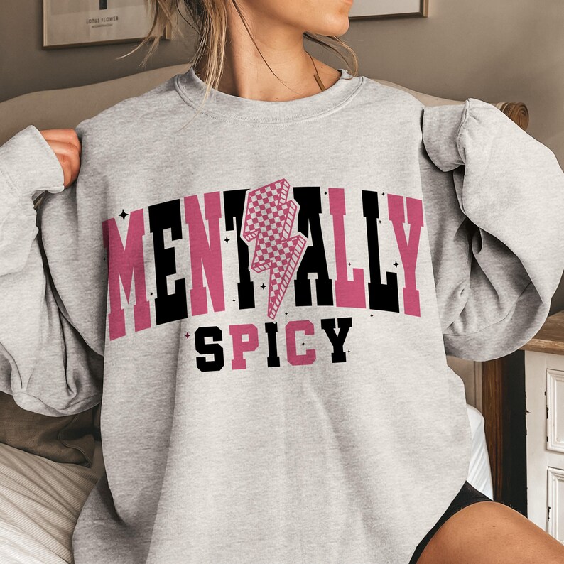 May include: A light gray sweatshirt with the words "MENTALLY SPICY" in a combination of pink and black block letters. A pink and white checkered lightning bolt graphic is centered above the word "SPICY". The sweatshirt has a crew neck and long sleeves.