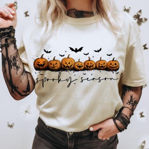 Op de afbeelding: Cr&egrave;mekleurig T-shirt met een rij uitgesneden oranje pompoenen en zwarte vleermuissilhouetten. De tekst "spooky season" staat onder de pompoenen, perfect voor Halloween.