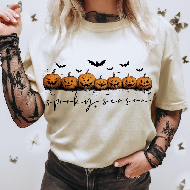 Op de afbeelding: Cr&egrave;mekleurig T-shirt met een rij uitgesneden oranje pompoenen en zwarte vleermuissilhouetten. De tekst "spooky season" staat onder de pompoenen, perfect voor Halloween.