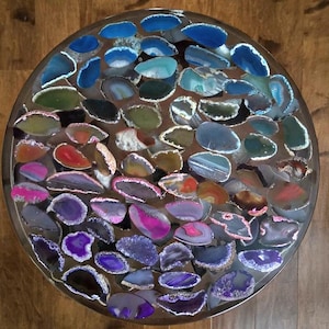 May include: A circular table top featuring a mosaic of polished agate stones in a spectrum of colours, including blue, green, red, purple, and pink. The stones are set in clear resin. A decorative element for interior design, suitable for a home.
