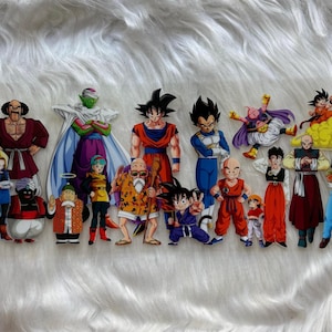 May include: A collection of colorful Dragon Ball Z character cutouts. The characters are in various poses and outfits, including Goku, Vegeta, and Piccolo. The cutouts are arranged on a white, fluffy surface.