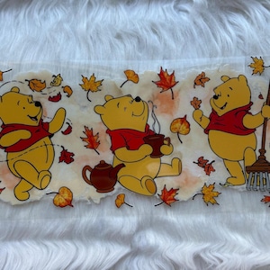 May include: A rectangular graphic featuring three cartoon bears in a fall scene. The bears are yellow with red shirts, surrounded by autumn leaves. One bear holds a broom, another a cup, and the third is waving.