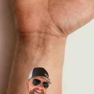 Pode incluir: Uma tatuagem tempor&aacute;ria com o rosto de um homem usando um bon&eacute; de beisebol preto e branco e &oacute;culos de sol. A tatuagem tamb&eacute;m inclui o nome "Brad" em escrita rosa e tr&ecirc;s cora&ccedil;&otilde;es rosas.