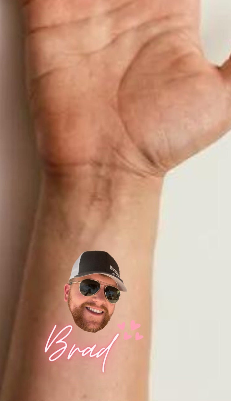 Pode incluir: Uma tatuagem tempor&aacute;ria com o rosto de um homem usando um bon&eacute; de beisebol preto e branco e &oacute;culos de sol. A tatuagem tamb&eacute;m inclui o nome "Brad" em escrita rosa e tr&ecirc;s cora&ccedil;&otilde;es rosas.