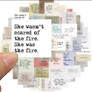 May include: A white square sticker with the text "She wasn't scared of the fire. She was the fire." is held in front of a collection of small, colorful paper notes with text. The background is blurred.