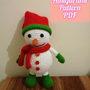 Puede incluir: Amigurumi de muñeco de nieve blanco tejido a crochet con un gorro rojo, ribete verde, bufanda roja y guantes verdes. El muñeco de nieve tiene una nariz de zanahoria, ojos negros y botones rojos.  Patrón Amigurumi PDF.