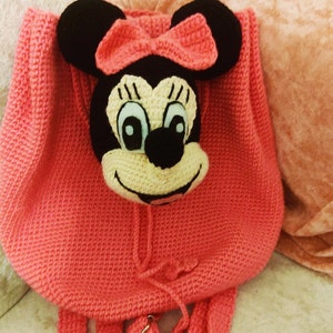 May include: A pink crocheted backpack with a Minnie Mouse face on the front. The backpack has two straps and a drawstring closure.