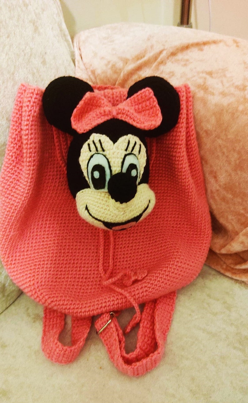 May include: A pink crocheted backpack with a Minnie Mouse face on the front. The backpack has two straps and a drawstring closure.