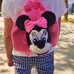 May include: A pink crocheted backpack with a Minnie Mouse face, black ears, and a pink bow. The backpack is being worn by a child.