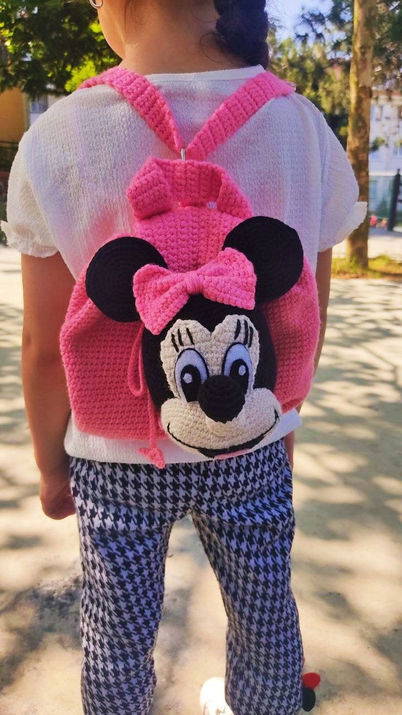 May include: A pink crocheted backpack with a Minnie Mouse face, black ears, and a pink bow. The backpack is being worn by a child.