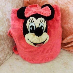 May include: A pink crocheted backpack with a Minnie Mouse face on the front. The backpack has a black nose, white eyes, and a black bow.