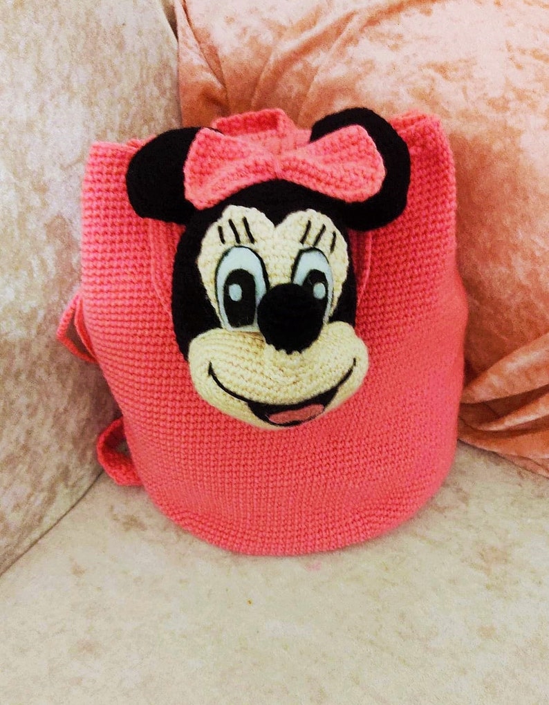 May include: A pink crocheted backpack with a Minnie Mouse face on the front. The backpack has a black nose, white eyes, and a black bow.