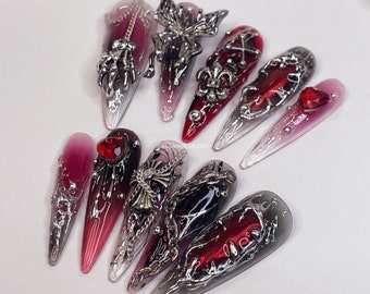 May include: A set of ten long, pointed, red and white ombre nail tips with silver and black embellishments. The embellishments include butterflies, crosses, and other gothic-inspired designs.