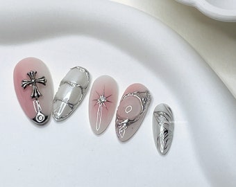 May include: Five artificial nails with various designs. One nail features a silver cross charm, another has a silver-lined white design, and a third has a starburst design. The other two nails have silver accents and marble-like patterns. The nails are painted in shades of pink, white, and gray.