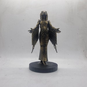 May include: A gold-colored figurine of a woman with wings and a bird-like head. The figurine is standing on a black circular base.