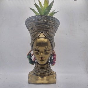 May include: A gold planter shaped like a woman's head with a green succulent plant growing out of the top. The planter has a braided hairstyle and beaded earrings.