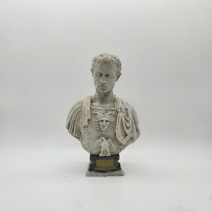 Puede incluir: Busto escultórico detallado de un hombre con pelo corto, vestido con una prenda tipo toga. La escultura es blanquecina con detalles intrincados, incluyendo una cara en el pecho y un águila. El busto está montado sobre una base oscura con una placa dorada.