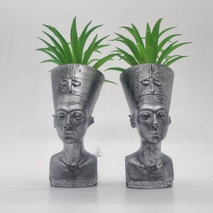 以下が含まれることがあります： エジプトのファラオの胸像の形をした銀色のプランターポット2つ。緑の植物が上から生えています。