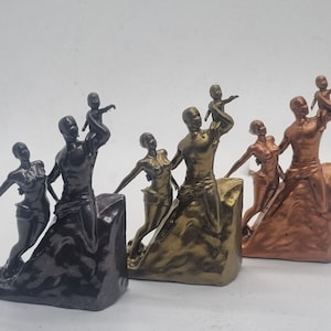 May include: Three different colored figurines of a family of four. The figurines are made of a dark brown, gold, and copper colored material. The family is standing on a rocky base. The father is holding a child in his arms, and the mother is holding a child in her arms. The children are looking up at the sky.