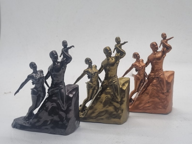 May include: Three different colored figurines of a family of four. The figurines are made of a dark brown, gold, and copper colored material. The family is standing on a rocky base. The father is holding a child in his arms, and the mother is holding a child in her arms. The children are looking up at the sky.