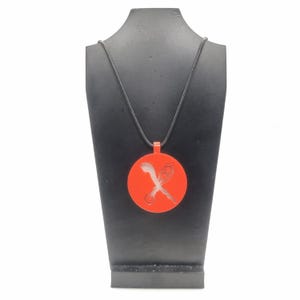 Puede incluir: Un collar con un colgante circular rojo y un cordón negro. El colgante presenta un diseño recortado de una espada estilizada y una luna creciente.