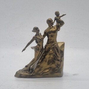 May include: A gold-toned metal sculpture of a family. The sculpture depicts a man holding a child in his arms, while a woman stands beside him. The sculpture is set on a base that resembles a rock.