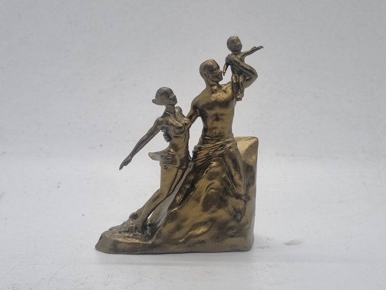 May include: A gold-toned metal sculpture of a family. The sculpture depicts a man holding a child in his arms, while a woman stands beside him. The sculpture is set on a base that resembles a rock.