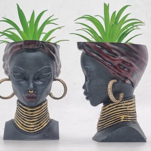 Puede incluir: Dos macetas de cerámica negra con forma de busto de mujer con un turbante rojo oscuro. Cada maceta tiene un collar y pendientes de oro. Las macetas están llenas de plantas verdes.