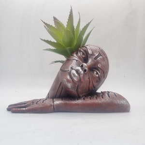 Puede incluir: Un macetero de cerámica marrón con forma de cabeza de mujer con una planta suculenta verde que crece en la parte superior.