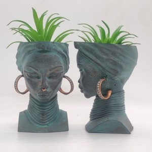 May include: Two green ceramic planter pots shaped like a woman's head with a plant growing out of the top. The pots have a textured surface and are decorated with gold hoop earrings.