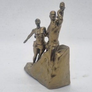 May include: A gold-colored figurine of a family, a man holding a child up in the air, a woman standing next to him, and a rocky base.