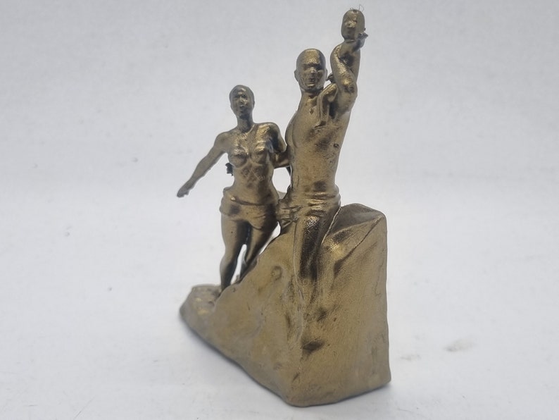 May include: A gold-colored figurine of a family, a man holding a child up in the air, a woman standing next to him, and a rocky base.