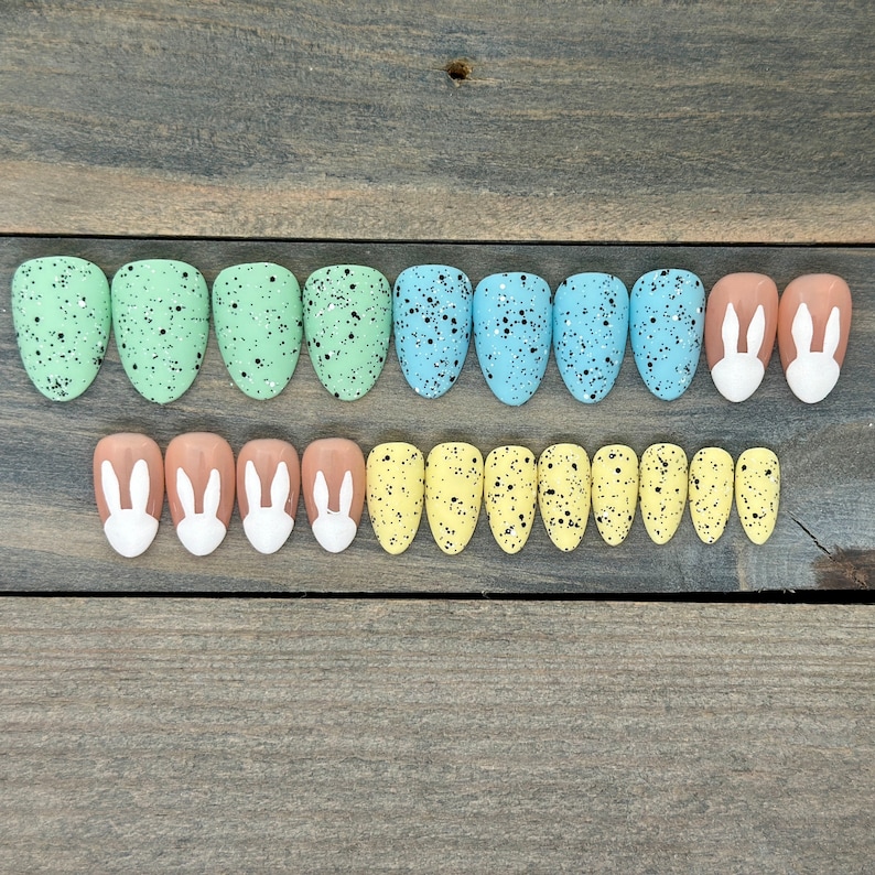 May include: A set of 18 false nails with Easter-themed designs. The nails are in three colours: light blue, light green, and light yellow. Some nails have a speckled design, while others have a bunny silhouette.