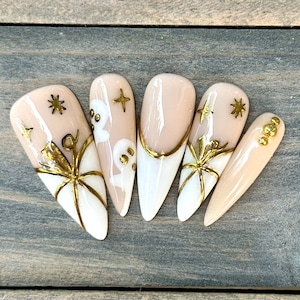 May include: Set of six long, almond-shaped false nails with a nude base and gold accents. Some nails feature a gold spiderweb design, others have gold stars, and one has a ghost design.