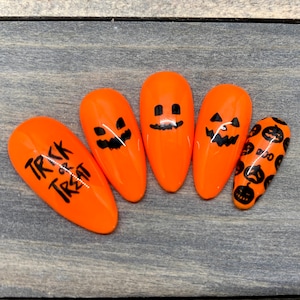 May include: Five orange Halloween-themed press-on nails. One nail has the text "Trick or Treat" written on it. Two nails have a jack-o-lantern face design. One nail has a jack-o-lantern face design with a small pumpkin on the side. One nail has a pattern of small jack-o-lantern faces.
