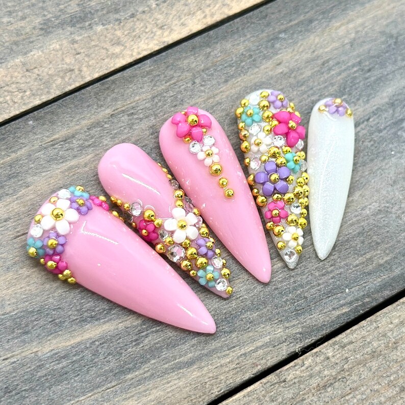 Puede incluir: Cinco u&ntilde;as postizas de color rosa en forma de stiletto con intrincados dise&ntilde;os florales. Cada u&ntilde;a est&aacute; adornada con peque&ntilde;as flores de colores y cuentas doradas.