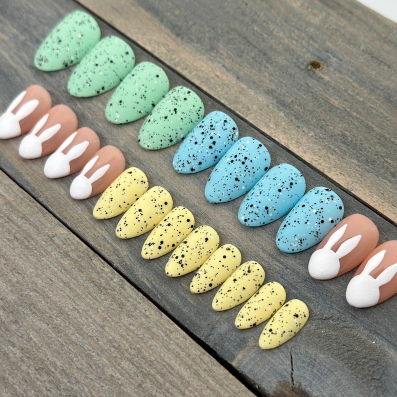 May include: A set of ten false nails with a speckled Easter egg design. The nails are in three colours: light blue, light green, and yellow. Each nail has a white bunny silhouette on the tip.