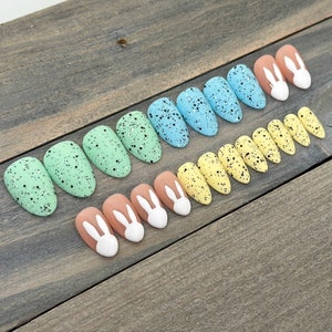 May include: Set of ten false nails with a speckled Easter egg design in shades of blue, green, and yellow. The tips of the nails feature a white bunny rabbit design.