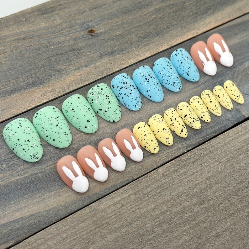May include: Set of ten false nails with a speckled Easter egg design in shades of blue, green, and yellow. The tips of the nails feature a white bunny rabbit design.