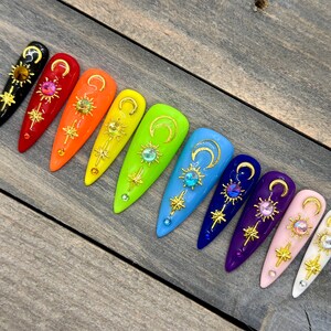 Peut inclure: Dix faux ongles longs et en forme d'amande avec un dégradé de couleurs arc-en-ciel. Chaque ongle est décoré d'un croissant de lune doré, d'une étoile dorée et d'un strass transparent.