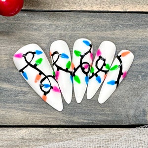 Peut inclure: Ongles artificiels blancs avec un motif de guirlande lumineuse. Chaque ongle présente un motif de lignes noires avec des lumières colorées bleues, vertes, roses et oranges. Les ongles sont en forme d'amande et disposés sur une surface en bois.