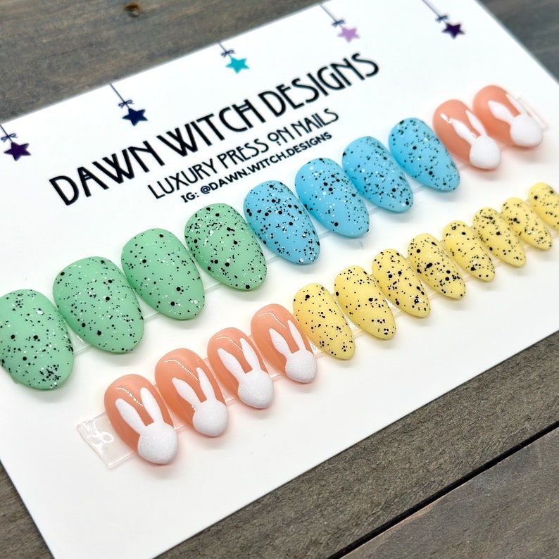 May include: A set of 10 press-on nails with an Easter theme. The nails are oval shaped and feature a speckled design in shades of green, blue, yellow, and peach. Each nail has a white bunny rabbit design.