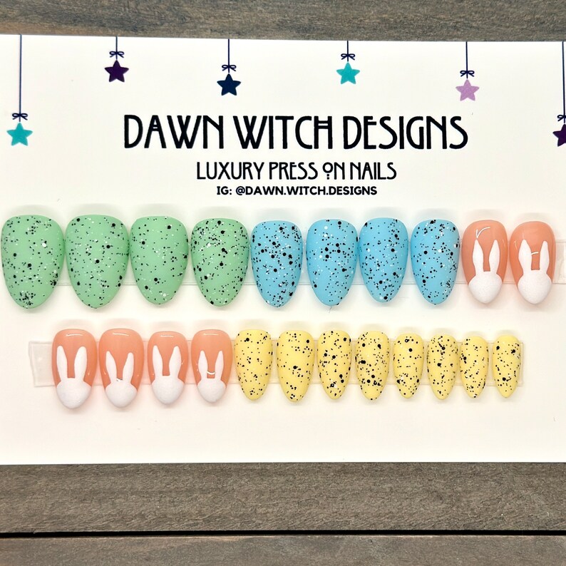 May include: A set of 12 press-on nails with an Easter theme. The nails are oval shaped and feature a speckled egg design in shades of green, blue, yellow, and peach. Some nails have a white bunny design. The nails are displayed on a white card with the text "DAWN WITCH DESIGNS" and "LUXURY PRESS ON NAILS" in black.
