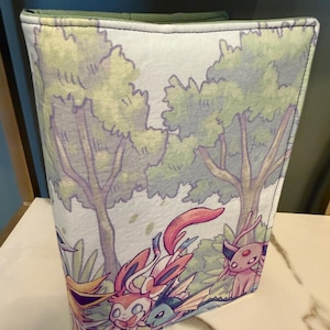 May include: A fabric sleeve featuring a colorful illustration of cartoon characters in a forest setting. The sleeve has a green trim and a white background. The characters are in shades of pink, blue, yellow, and purple.