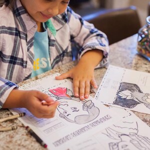 Puede incluir: Un ni&ntilde;o colorea un dibujo en blanco y negro de una figura hist&oacute;rica con un cray&oacute;n rojo. El ni&ntilde;o lleva una camisa a cuadros y una camiseta azul. Hay crayones y un tarro de crayones sobre la mesa.